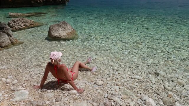 Woman in pebble beach with feet in serene Adriatic Sea. Enjoying a summer vacation in a hidden cove surrounded by natural rocky cliffs under bright blue sky in Cres Island of Croatia