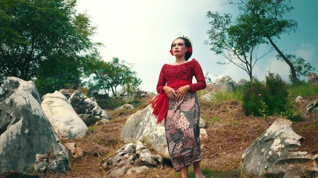 An elegant Indonesian woman in traditional red clothing posing confidently among scattered white rocks and green trees under the sunlight.
