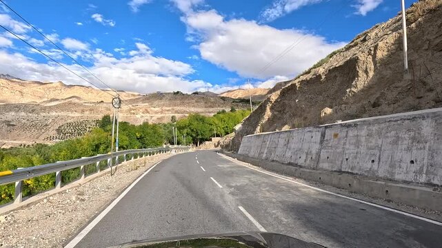 A vehicle curves past an eroded cliff with a green valley and village with orchards visible below and barren desert mountai - Spiti Valley Himachal Pradesh India | Himalayan road trip GoPro dashcam 4K