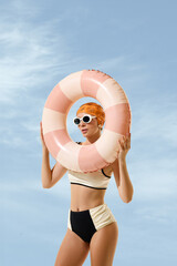 Playful woman framing face with pink swim ring in sky themed studio wearing white sunglasses. Summer fun, travel ads, eyewear fashion concept. © Lustre Art Group 