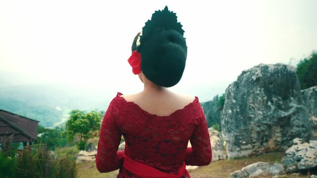 Close-up back shot of a woman wearing a traditional Javanese bun and red lace Kebaya standing still and gazing at the green landscape from a rocky hill.