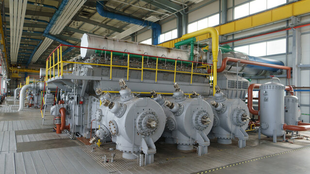 View of a large industrial gas compressor engine inside a power plant facility with complex piping systems, yellow safety railings, and metal flooring in Latvia.