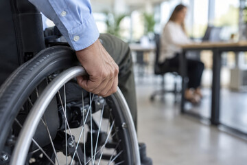 Obraz premium A person using a wheelchair is visible in a modern office space. Another individual is sitting at a desk, focused on work activities, creating a collaborative atmosphere