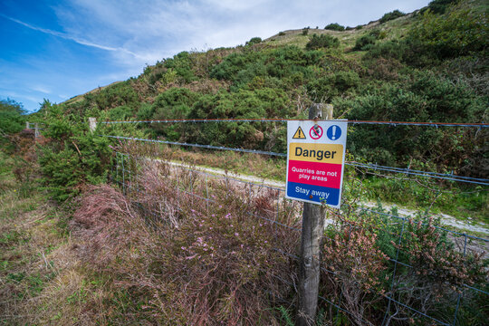 View of a danger warning sign for the Blackpool Clay pits attached to a barbed wire fence with a grassy hillside in the background in St Austell, Cornwall, United Kingdom.