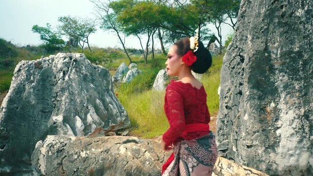 A wide shot of an Indonesian dancer wearing traditional attire walking through a jagged limestone garden and sitting down to rest on a flat stone surface.