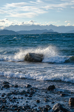View of waves crashing against a large rock on the pebbled shore of Nahuel Huapi Lake with distant mountains in San Carlos de Bariloche, Rio Negro, Argentina.