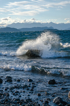 View of waves crashing against a rock on the rocky shore of Nahuel Huapi Lake with distant mountains under a blue sky in San Carlos de Bariloche, Rio Negro, Argentina.