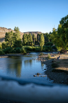 View of people relaxing on the banks of the Rio Chimehuin with lush trees and hills under a clear blue sky in Junin de los Andes, Neuquen, Argentina.