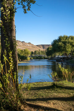 View of Rio Chimehuin with a bridge in the background, lush trees along the riverbanks, and a person fishing in the water in Junin de los Andes, Neuquen, Argentina.