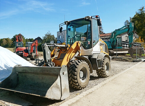 View of a yellow wheel loader parked at a construction site with an excavator in the background under a clear blue sky in Garching, Bavaria, Germany.