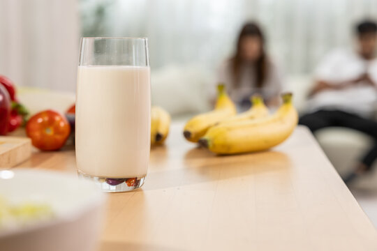 Milk glass on kitchen table with banana and fresh fruit, blurred family background creating warm breakfast scene