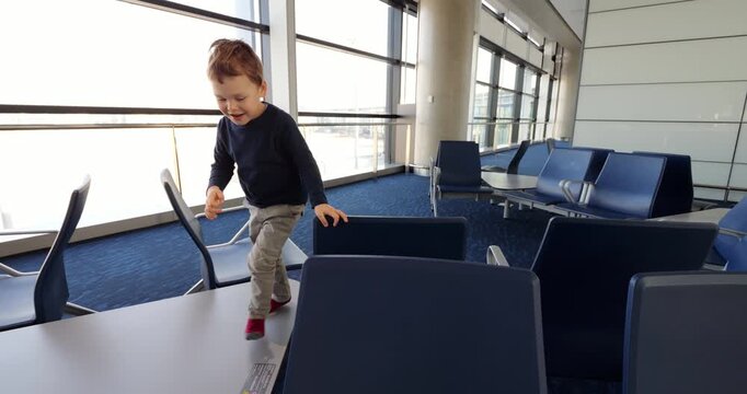 Walking across empty passenger seats and armrests, energetic toddler climbs and balances along geometric cluster of lounge chairs inside vacant airport terminal. Brave little child seeks recreation