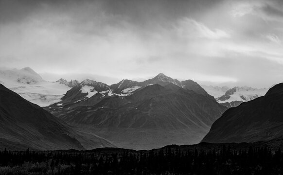 View of the rugged, snow-capped mountains of the Alaska Range under a dramatic, cloudy sky with a dense evergreen forest in the foreground in Paxson, Alaska, United States.