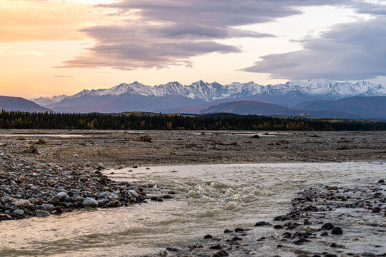 View of the Gerstle River flowing through a rocky landscape with snow-capped mountains in the background under a soft sunset sky in Deltana, Alaska, United States.