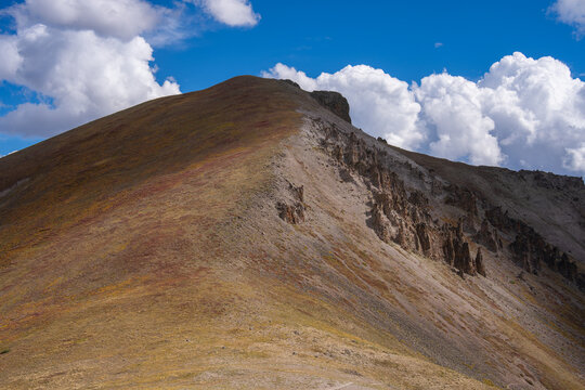 View of the rugged slopes and rocky formations of San Luis Peak under a bright blue sky with scattered clouds in Creede, Colorado, United States.