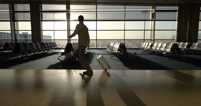 Running through spacious airport terminal lounge, father pulls rolling suitcase while his little son trails bit behind following his steps. Late passenger dad glances back to encourage son to run fast