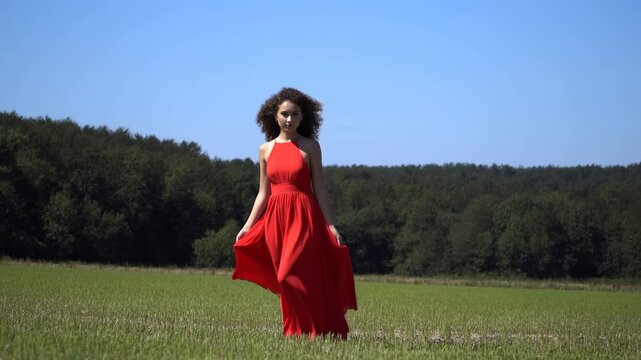 Woman walks toward camera through sunlit green field in flowing red maxi dress, holding skirt edges as fabric moves gently against distant forest and clear blue sky