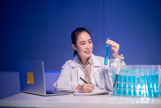 Asian Female Scientist Examining Blue Liquid in Test Tube at Laboratory Desk with Laptop and Samples