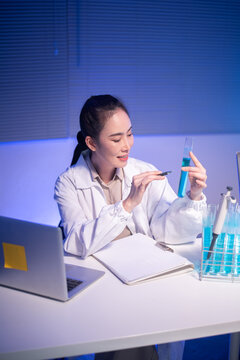 Asian Female Scientist Examining Blue Liquid in Test Tube at Laboratory Desk with Laptop and Glassware