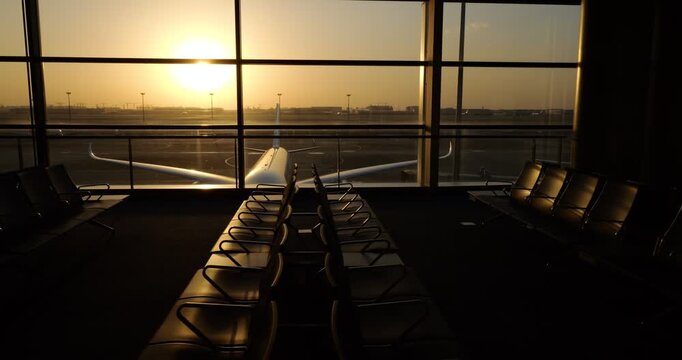 Gleaming in golden hour sunlight, vacant terminal waiting area features rows of empty seats, camera looking out through large panoramic windows toward commercial jet airliner parked near terminal