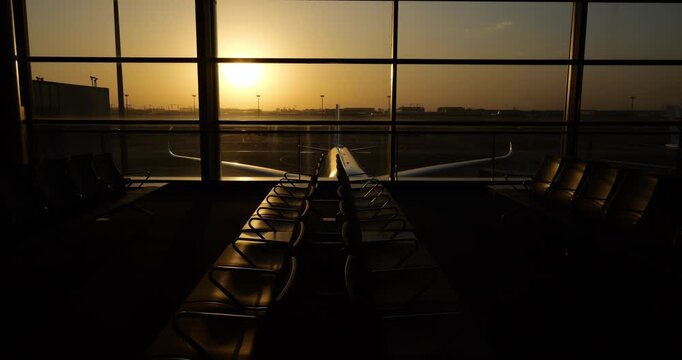 Revealing empty terminal lounge seats, camera pans slowly across deserted boarding waiting area at golden hour. Large panoramic glass windows overlook commercial airliner parked near terminal