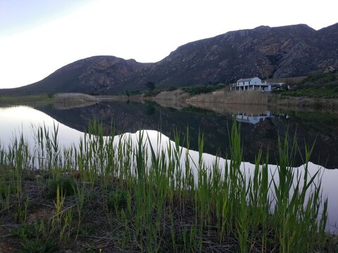View of a serene pond reflecting the rugged mountains and a house in the landscape of Montagu, Western Cape, South Africa.