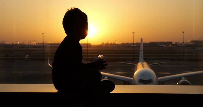 Sitting on panoramic window sill, toddler boy watches large commercial airplane parked on apron tarmac right outside. Raising head curiously to track sudden motion outside, child adjusts posture