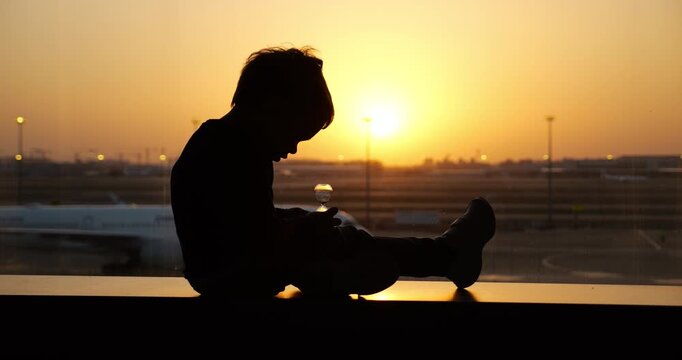 Dreaming while watching last sand grains fall, silhouetted toddler sits quietly on airport terminal window sill. Blurred background with airplane parked on tarmac and bright golden sunset.
