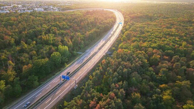 Curved divided highway through autumn forest in golden light with moving vehicles, overhead directional signs, and central barrier near suburban edge at sunset