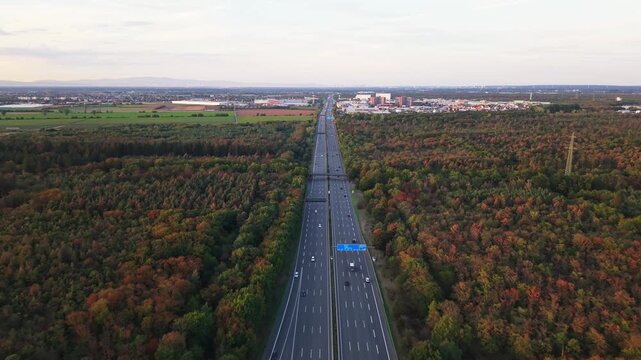 High aerial view of divided multi-lane highway through autumn forest, leading toward industrial town and farmland with scattered vehicles and power lines in warm evening light