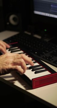 Older musician plays digital piano keyboard in home music studio. Hands create melody beside laptop and studio monitor. Warm close-up shows creativity, recording, and music production.