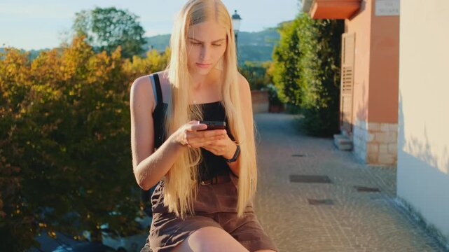 A young blonde woman in a dark camisole dress sits on a stone bench outdoors, focused on her smartphone beside Mediterranean-style architecture during daytime.