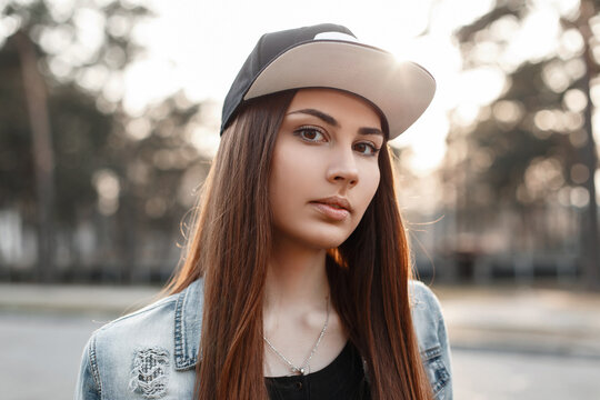 Close-up portrait of a beautiful hipster girl in a black baseball cap and torn blue jeans jacket at sunset