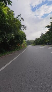 Scenic winding roadway through lush green forest with chevron warning signs and concrete barriers under a partly cloudy sky, showing a peaceful tropical road landscape.