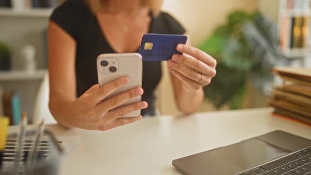 Woman holds smartphone and creditcard with hands at a home office work desk; online payment confidence.