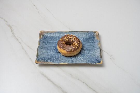 Chocolate-glazed mini donut sprinkled with crushed nut pieces on a rectangular blue ceramic plate over white marble