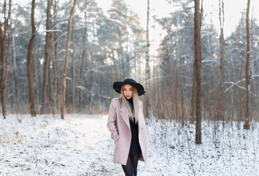 Pretty fashionable woman in black hat and pink coat in winter amazing park with woods at sunset. Beauty winter girl