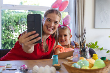 Obraz premium Mother and child in bunny ears using phone painting Easter eggs with paints, brushes at home