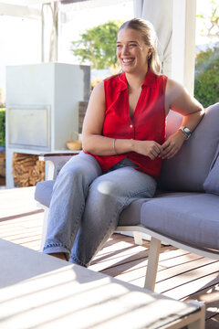 Woman in red blouse sitting on gray sofa under pergola, checking smartwatch and smiling, fireplace