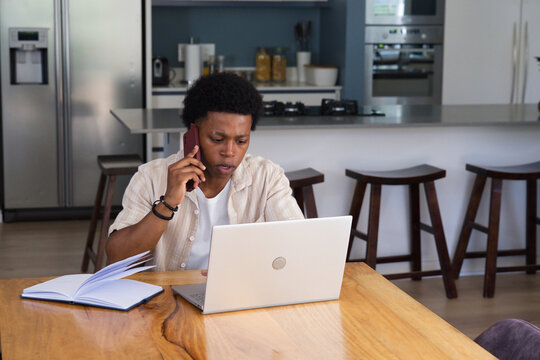 African man wearing light beige shirt holding smartphone and working on laptop at dining table