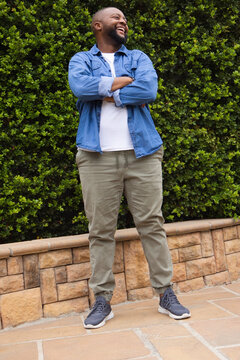 African American man standing in courtyard laughing, looking up left, wearing denim shirt, hedge