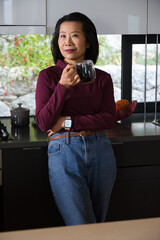 Mature Asian woman standing in modern kitchen holding glass mug wearing burgundy top and smartwatch © WavebreakMediaMicro