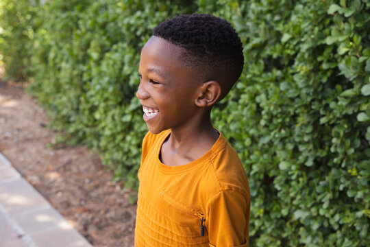 African American child standing on pavement by green hedge soil orange zip shirt smiling