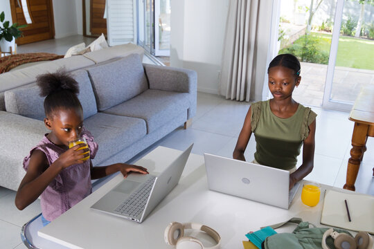 African female siblings child and youth using two silver laptops and drinking orange juice at home