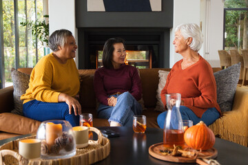 Obraz premium Diverse female friends in sweaters and jeans chatting and sipping amber drinks on sofa by fireplace