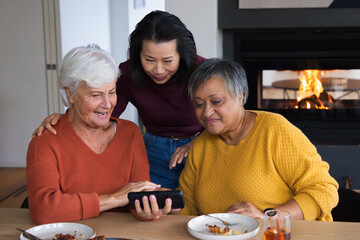 Obraz premium Diverse female friends leaning around wooden dining table at home, sharing smartphone and plates