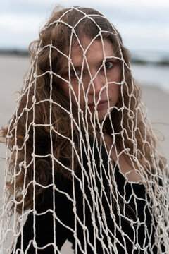 Woman with fishing net draped over her hair and shoulders. She faces the camera in a black neckline against a softly blurred strip of shore and sea.