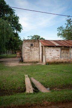 View of a rustic brick house with a weathered corrugated metal roof set in a grassy rural landscape with wooden planks crossing a small ditch in Guaviravi, Corrientes Province, Argentina.
