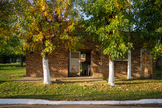 View of a rustic brick house with white-painted tree trunks in the foreground and autumn foliage in Guaviravi, Corrientes Province, Argentina.