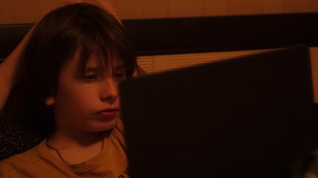 A teenage boy sits in front of a laptop screen in a dark room and watches online content or studies. A young child spends the evening at home focused on the computer monitor